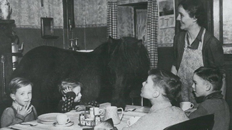 Black and white photo of children sitting around a dinner table waiting to be served their food and a lady in an apron standing. A black pony is eating from the table too!