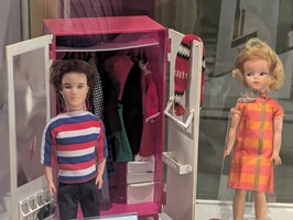 Two dolls are displayed, one wearing a striped shirt standing beside an open pink wardrobe filled with clothes. The other doll is dressed in an orange checkered outfit and stands slightly to the right. The background is blurred.