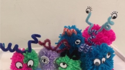 Colorful pom pom creatures with googly eyes and pipe cleaner antennas on a white background. They are in shades of pink, blue, green, and purple, with various expressions and eye designs.