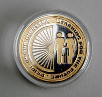 A round coin in a clear case features a gold and silver design with silhouettes of two adults and a child. Text around the edge reads, "Pride in our industry. Learning for the future.
