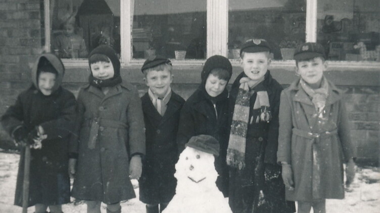 Six children in winter coats and hats stand in the snow, smiling beside a snowman with a hat and scarf. They are in front of a building with large windows. One child holds a small shovel.
