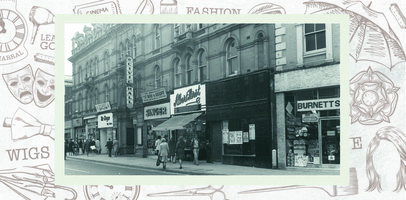 Civic Hall signage above its entrance and shopfronts for Mr Wayne, Singer, Albert Hirst and Burnetts. A sign for a performance at the Civic Hall called Ros and Geoff is visible above the Singer shop.