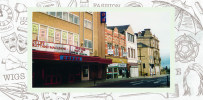 Odeon cinema with film advertisements for Bugs Life, Plunkett and MacLeane, Urban Legend and Mighty Joe. Pricing posters in first floor windows. Next to the cinema is a panini shop, followed by a charity shop.