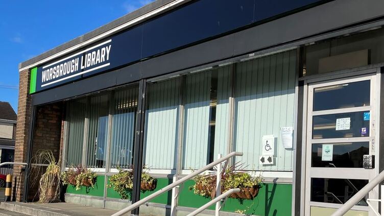 Library frontage with black sign, hanging plants and blinds in the windows with steps up to the entrance door.