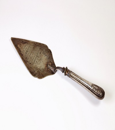 A vintage, ornately engraved silver cake server with a tapered, pointed blade and a decorative handle, resting on a white background.
