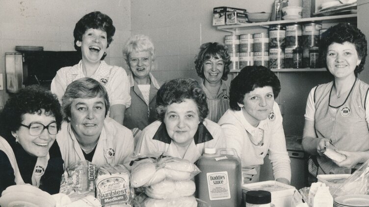 Women posing for a photograph in one of the soup kitchens, they are surrounded by bread, sauce bottles etc