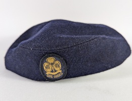 A navy blue beret with an embroidered patch featuring the Girl Guides logo. The patch shows a clover-like symbol with the letters "GG" and the words "Girl Guides" in yellow. The beret is positioned on a plain white surface.