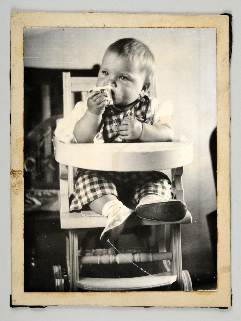 A black-and-white vintage photo of a baby sitting in a wooden high chair, wearing plaid overalls and a white shirt. The baby is holding and eating a piece of bread, with a thoughtful expression. The background is blurred, focusing on the child.