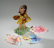 Vintage paper doll standing on a floral base, dressed in a yellow coat while holding a basket and hat. Three additional outfits lie around: a pink dress, a blue dress, and a green dress, each with detailed decorations.