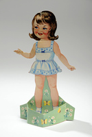 A vintage paper doll of a young girl with brown hair, wearing a light blue dress with floral patterns. She stands inside a green base decorated with daisies and butterflies on a white background.