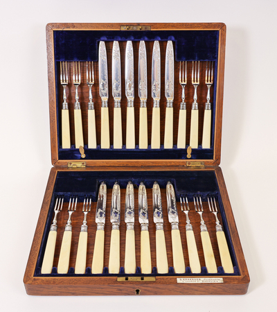 A wooden box with a blue velvet lining displays a set of cutlery, including knives and forks with cream-colored handles, arranged neatly. The lid holds larger utensils, while the bottom section contains smaller ones.