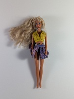 A doll with long blonde hair is wearing a yellow sleeveless top and a purple patterned skirt with blue details. The background is plain white.