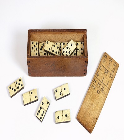 A set of vintage dominoes scattered on a white surface, with some pieces placed next to an open wooden box. A wooden tally sheet with markings is beside the box, suggesting game scores. The dominoes are cream-colored with black dots.