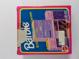 The image shows a pink and blue Barbie-branded toy packaging of an "Entertainment Center." The box features graphics of toy stereo equipment. A section of the packaging is torn, revealing a bit of the contents inside.