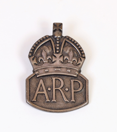 A bronze badge featuring a crown design above the letters "A.R.P." against a white background. The badge has a curved top and flat bottom, with the letters prominently embossed.