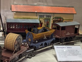A model railway scene featuring a train station with a ticket office and bookstall. Several train cars, including a coal car and spool car, are on the tracks. A sign reads "Sampler, 1863" with text about school samplers made by girls.