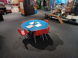 A small round blue craft table with red chairs in the main Gallery with paper, pens and trails to do