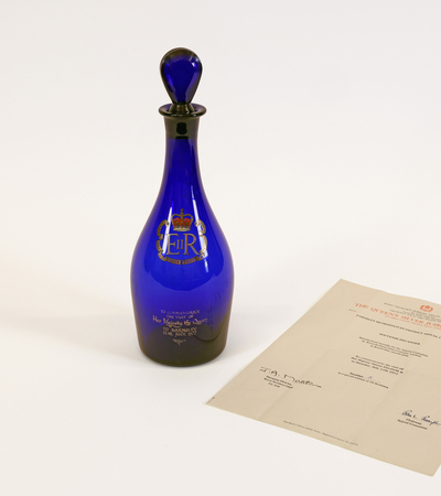 A cobalt blue glass bottle with a royal insignia and a round stopper sits beside an official certificate on a plain white background. The certificate has signatures and text, indicating its significance.
