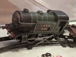 A model steam locomotive with the label "LNER 2162" is displayed on a track. The engine is dark green with light green lining and features intricate detailing, including visible wheels and a connector to another model piece in the background.