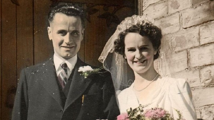 Man in dark suit and lady in white wedding dress in front of a church door.