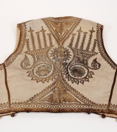 Ornate traditional vest with intricate embroidery in gold and blue hues on a beige background. The design features symmetrical patterns and geometric shapes, with buttons along the bottom edge. The vest is displayed on a plain white surface.