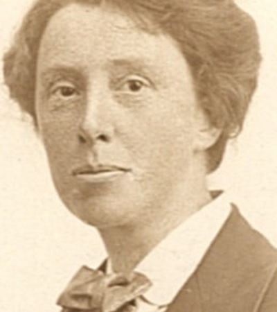 A sepia-toned portrait of a person with short, neatly styled hair, wearing a jacket, white shirt, and bow tie, looking directly at the camera with a neutral expression.