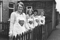 Four women in aprons standing outside resting against a wooden fence